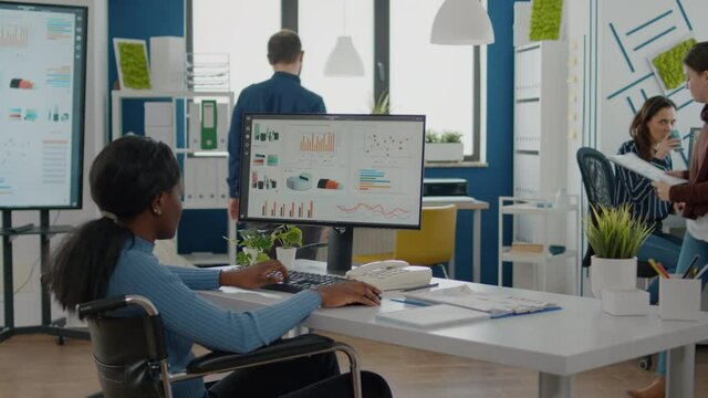 Businesswoman coach leader speaking with african employee with disabilities checking business report explaning financial graphs on computer, black paralysed woman in wheelchair and coworker discussing