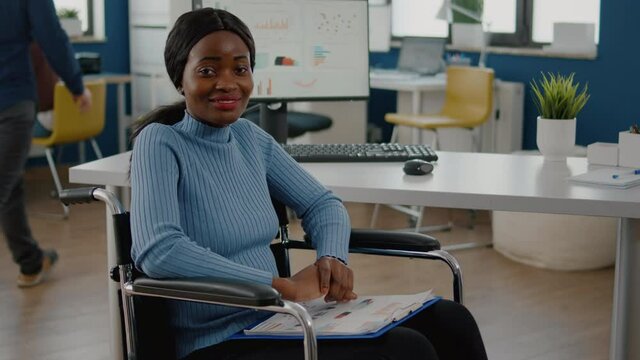 Immobilized paralized handicapped african woman employee looking at camera smiling sitting in wheelchair in business office room working at financial project with team. Businesswoman with disabilities