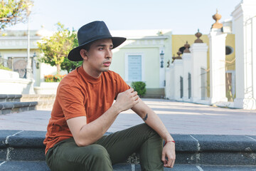 Young man wearing hat, sitting in a garden in thoughtful pose.
