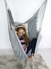 Little girl with headphones enjoying music in a hammock. Portrait on white wall inside.