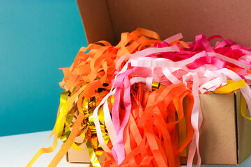 Paper tassel garlands in a cardboard box on the table against blue wall. Colorful sparkling holiday paper ribbons. Celebration curly mixed golden, yellow, pink tapes. Birthday party, festive, fun. 