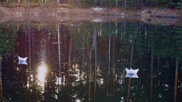 Paper boats peacefully sail on the water surface in the rays of the setting sun breaking through the trees and reflecting in the water along with coniferous trees. Origami boat.