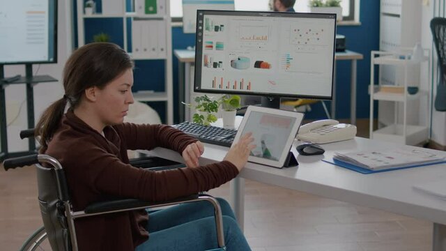 Invalid paralysed handicapped woman entrepreneur using computer and tablet in same time checking statistic datas working in business start-up office in wheelchair. Busy multitasking employee analysing