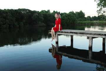 Fototapeta na wymiar A young blonde woman in a red dress sits on the footbridge near the village pond. The romance of loneliness. Loneliness at sunset