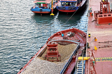 Loading bulk cargo of limestone from barges into a bulk carrier by using ship cranes and grabs at...