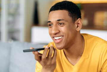 Positive attractive mixed race hispanic guy, sitting on sofa, wearing t-shirt, using smartphone,...