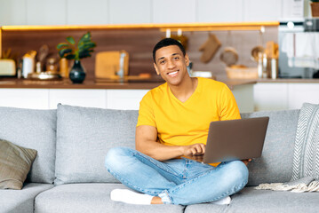 Satisfied handsome young latino man in casual clothes, sitting on the couch at home in the living...