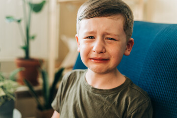 Close up portrait of crying little kid boy, sitting on couch indoors at home. Upset child