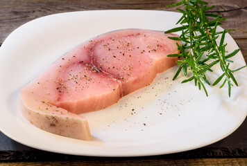 Raw Swordfish fillet covered with black pepper and with rosemary sprigs on white plate.