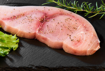 Raw Swordfish fillet covered with black pepper and with rosemary sprigs on black slate plate. - obrazy, fototapety, plakaty