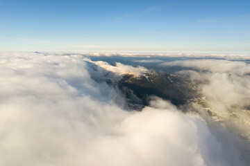 Obraz premium Aerial view from above of white puffy clouds covering snowy mountain tops in bright sunny day.