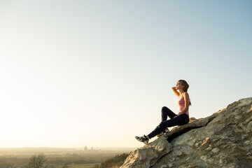 Woman hiker sitting on a steep big rock enjoying warm summer day. Young female climber resting during sports activity in nature. Active recreation in nature concept.