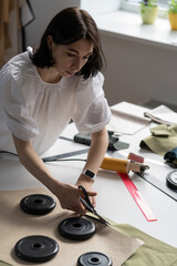 Fototapeta na wymiar Woman professional seamstress hold scissors cut material for sewing on working table in atelier. Tailor girl prepare cloth use weights and patterns. Young fashion designer or freelance stylist at work