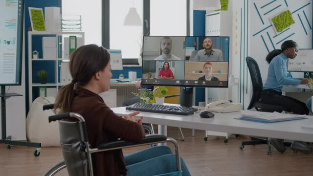Handicapped amputee paralysed project manager talking with team during video conference using videocall sitting in wheelchair in startup business office. Corporate partner discussing in online meeting