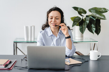 Confident mature middle-aged businesswoman in headset speaking on conference call on laptop, female...