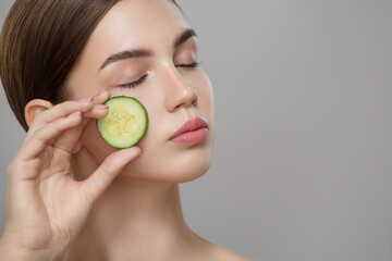 Organic cosmetics concept. A young girl with clean skin holds cucumber slice near her face. Gray background.
