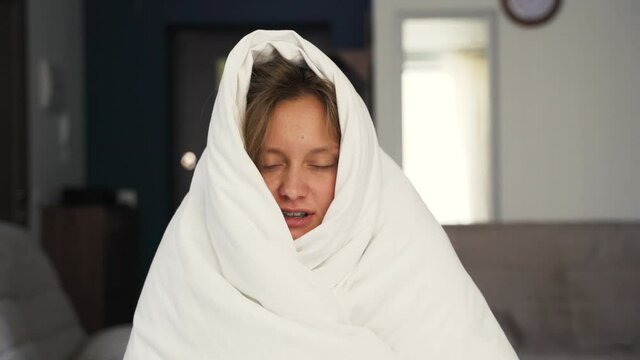Symptoms of coronavirus. A sick young woman, feeling cold fever, freezes without central heating, feeling cold at home. Covered with a warm white blanket. Shivering, sitting on the couch, stressed out