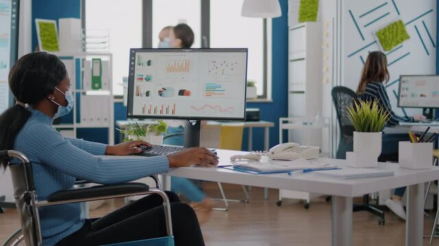 African disabled paralyzed businesswoman with protection mask writing financial reports on computer in busy corporate company office working with cowerkers respecting social distance against pandemic