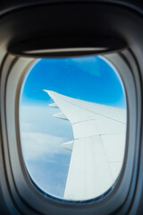 Fototapeta na wymiar airplane wing through the window porthole on background of blue sky and white clouds, mood cold color toning, vertical travel content