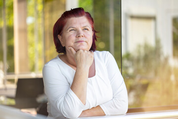 Depressed senior woman feels sad at home. The old woman is looking out the window. Depressed thoughtful lonely lady standing alone and looking out the window.
