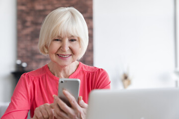 Smiling old elderly senior woman holding smartphone and using laptop at home office. Successful old woman working at home typing messages on freelance job.