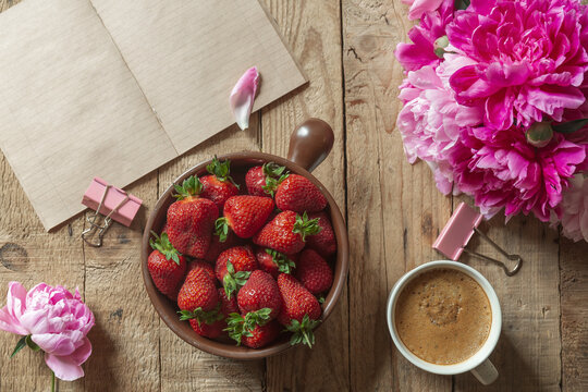 Ripe strawberries in clay bowl, bouquet of peonies, cup of coffee and vintage notebook on a wooden table. Top view, copy space .