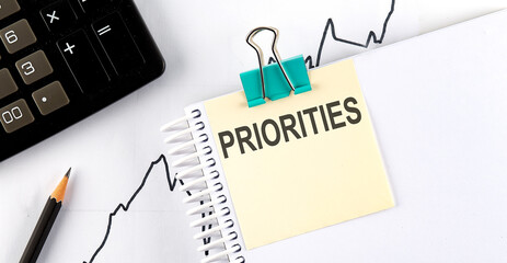 Notepad with text PRIORITIES on a white background, near calculator and office supplies. Business...