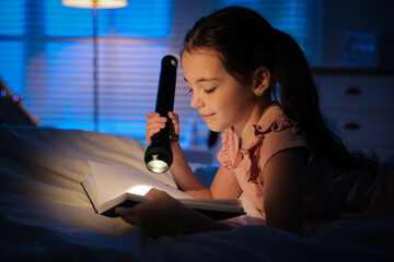Little girl with flashlight reading fairy tale in dark bedroom