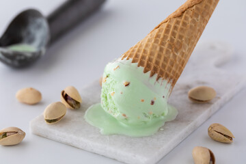 A tipped over pistachio ice cream cone on a small marble slab surrounded by pistachios and a metal scooper in behind.