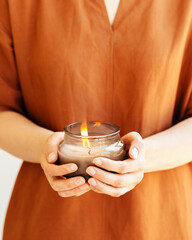Woman hand holds lit scented candle in glass jar with natural ingredients on terracotta
