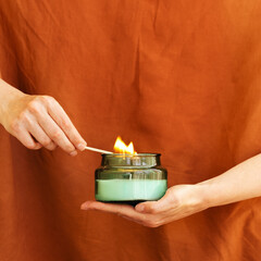 Womans hand uses match to light aromatic candle in recycled glass jar on terracotta color