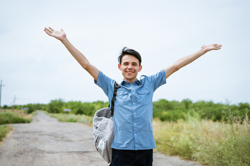 Fototapeta na wymiar Happy young man posing with raised arms, standing on the road and looking into the distance. Happy guy in a blue shirt with a backpack. Free student enjoys vacation