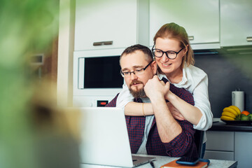 Adult married couple in cozy home interior. Husband and wife look at the laptop screen, discussing...