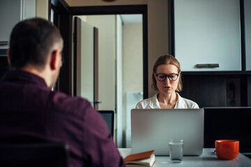 Adult young married couple sitting at a table in the kitchen, remotely working together on laptops, discussing business and smiling. Focus on handsome woman with glasses. Man sits back to the camera.