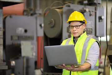 Asian engineering manager in safety hard hat and reflective cloth is inspecting inside the factory using digital laptop