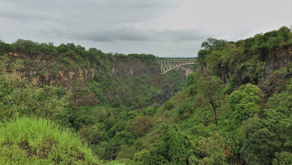 An arched bridge runs over a deep gorge. Lush tropical vegetation on the steep slopes and at the bottom. Cloudy sky. Zambia
