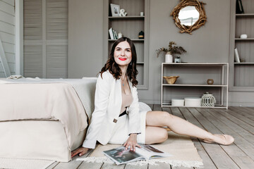 A young woman in a white suit, skirt in the house is sitting on the floor near the bed flipping through a magazine and drinking tea.