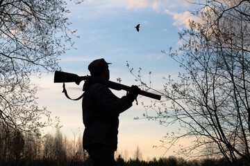 a hunter with a rifle on his shoulder looks at a flying woodcock late at night - obrazy, fototapety, plakaty