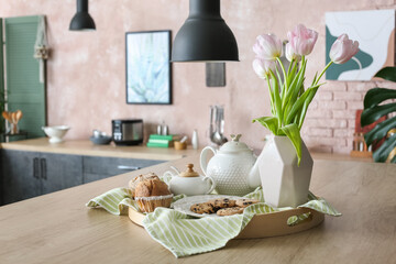 Tray with tasty muffins, teapot and vase on table in kitchen