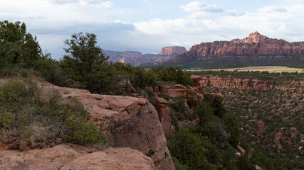Clouds drift by in the sky above the red sandstone rim of a mesa in Southern Utah. Juniper trees and small bushes grow on the top and sides of the cliffs.