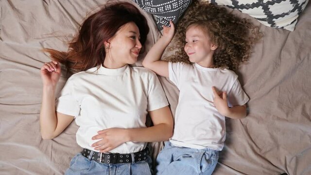 Relaxed brunette mother and curly haired preschooler daughter lie on large grey bed and kiss near black white pillows close upper view
