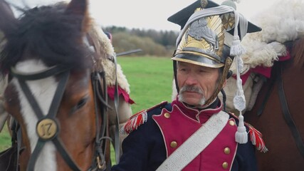 Portrait of a adult male hussar in medieval costume, a soldier standing next to a horse in a field, close-up, 4k slow motion.
