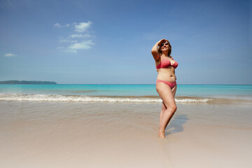 Fototapeta na wymiar Full body of smiling overweight woman covering face from sunlight while standing on sandy beach near waving sea