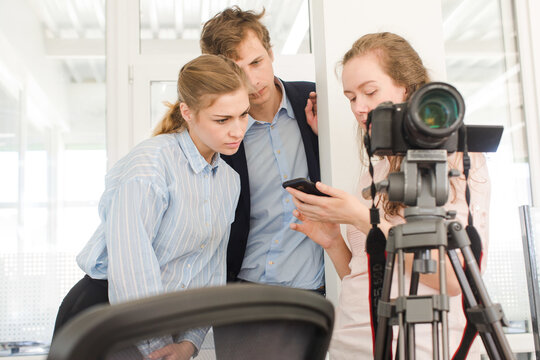 Group of young creative coworkers standing near tripod with professional camera and discussing photos on mobile phone while working together in modern workplace