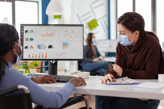 Immobilized handicapped invalid african entrepreneur working with colleague in new normal business office during covid19 pandemic wearing face masks, checking graphs calculating profit on computer.