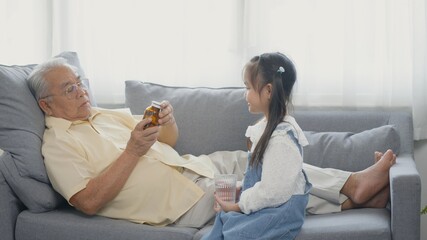 Obraz na płótnie Canvas Asian granddaughter brought medicine and water for Grandpa to eat on the sofa in the lounge, Senior old man taking pills medicine time, granddaughter take care older with love