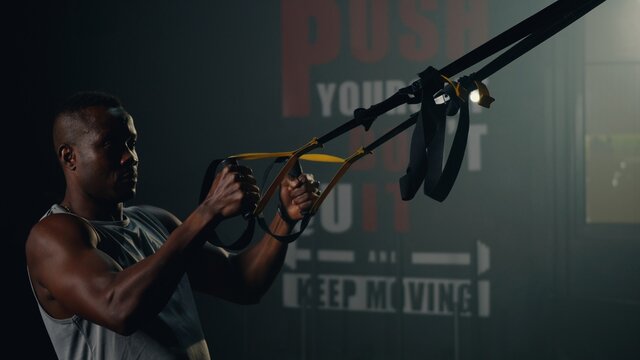Black muscular man during exercise training workout bodyweight with suspension straps TRX pull ups training arms at stylish empty gym center fitness GYM, workout healthy lifestyle sport concept