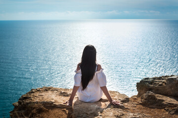 Fototapeta na wymiar girl sitting on a rock. rejoices in freedom. cliff in the sea. horizon of the ocean. back view. copy space