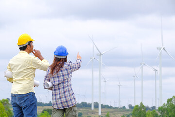 An engineer stands holding a blueprint and looks at a wind turbine project to generate electricity and check wind direction.