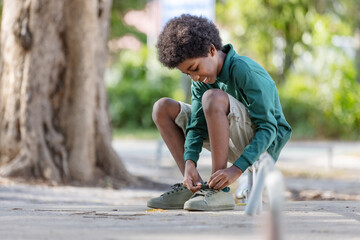 A black curly haired African boy sits on an iron railing with shoelaces at outdoor.